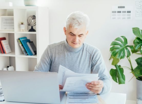 Middle aged businessman reading paper documents and using laptop while working at home office.