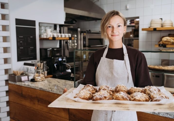 Welcoming,Female,Baker,Holding,Freshly,Baked,Almond,Croissants,In,Background